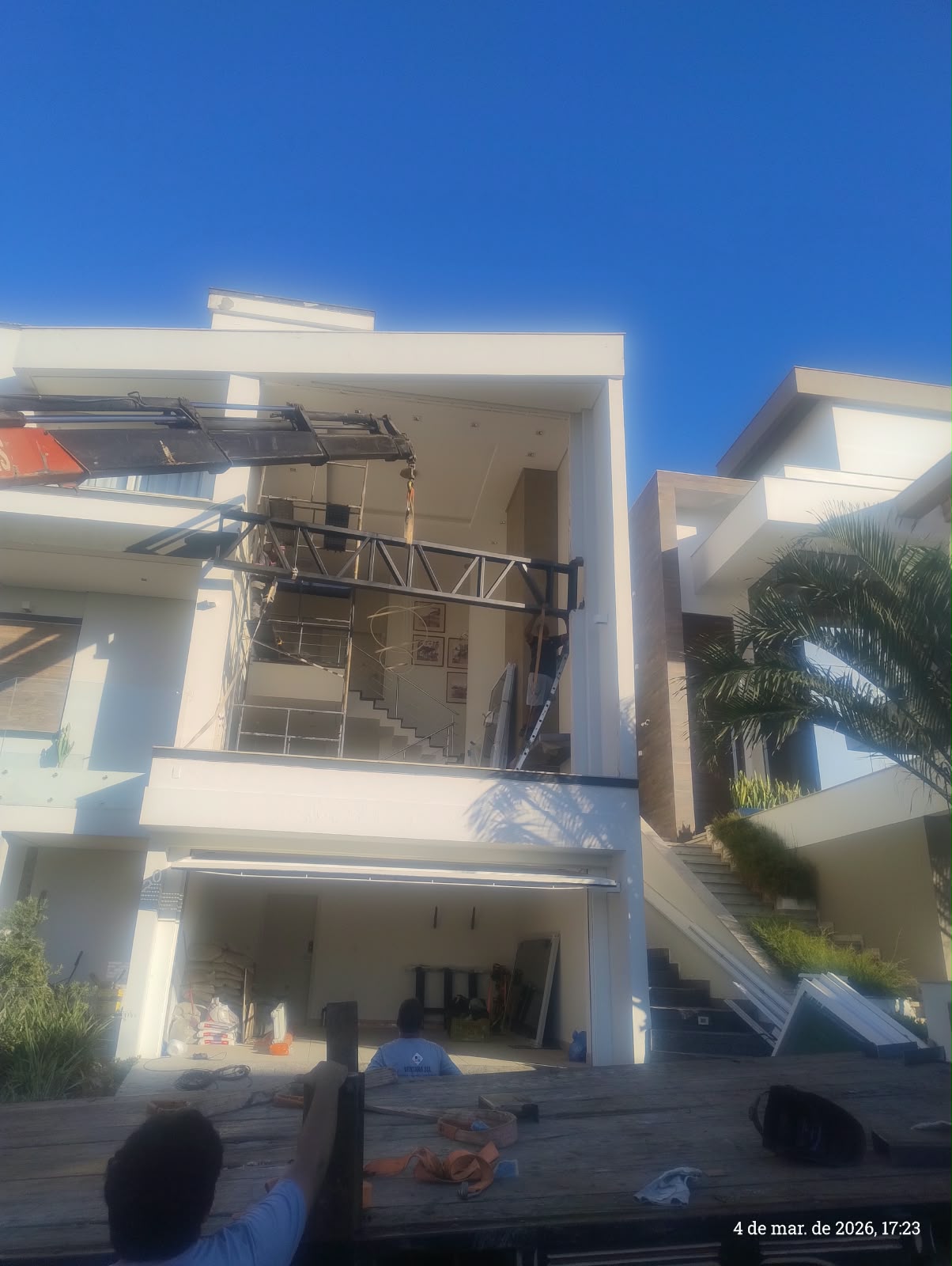 Equipamento de içamento movimentando uma estrutura metálica de reforço diante do vão superior da residência em obra.